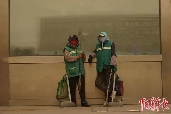 4月19日，内蒙古包头，沙尘暴来时，包头火车站的两名清洁工在墙边躲避风沙。李强 / 摄